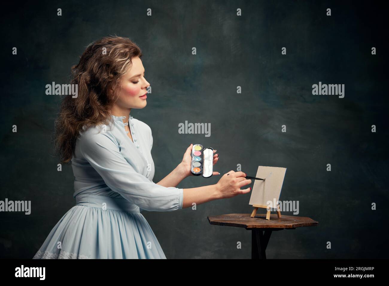 Künstler. Porträt einer charmanten aristokratischen Frau, die ein blaues historisches Kleid trägt, die Lidschatten in der Hand hält und sie auf eine kleine Leinwand zeichnet. Stockfoto