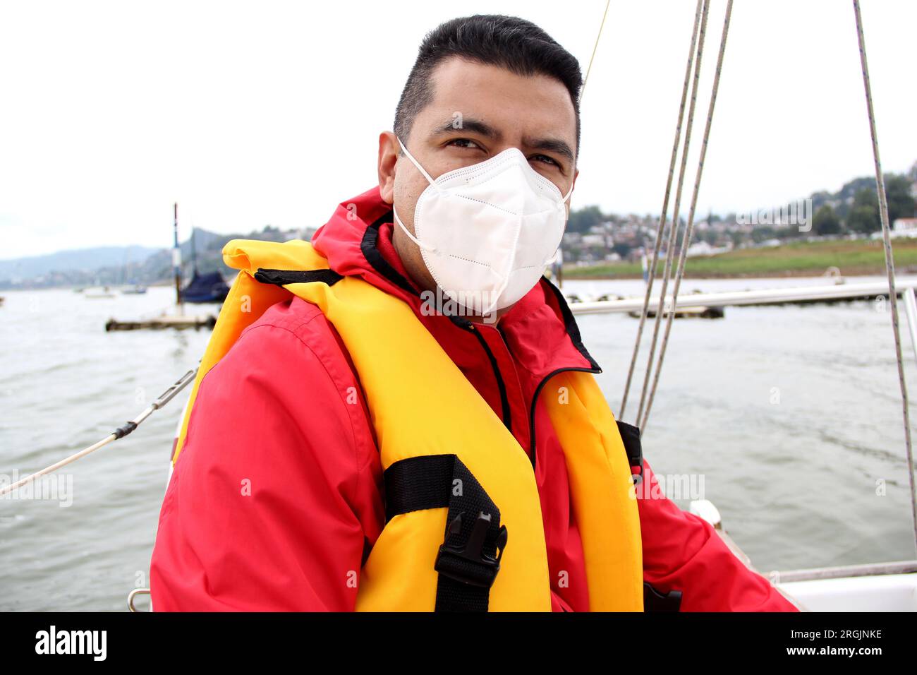 Ein erwachsener Latino-Mann fährt ein Segelboot auf dem See mit Windjacke und Schwimmweste, trägt Gesichtsmasken in der neuen Normalität aufgrund der Covid-19-Pandemie Stockfoto