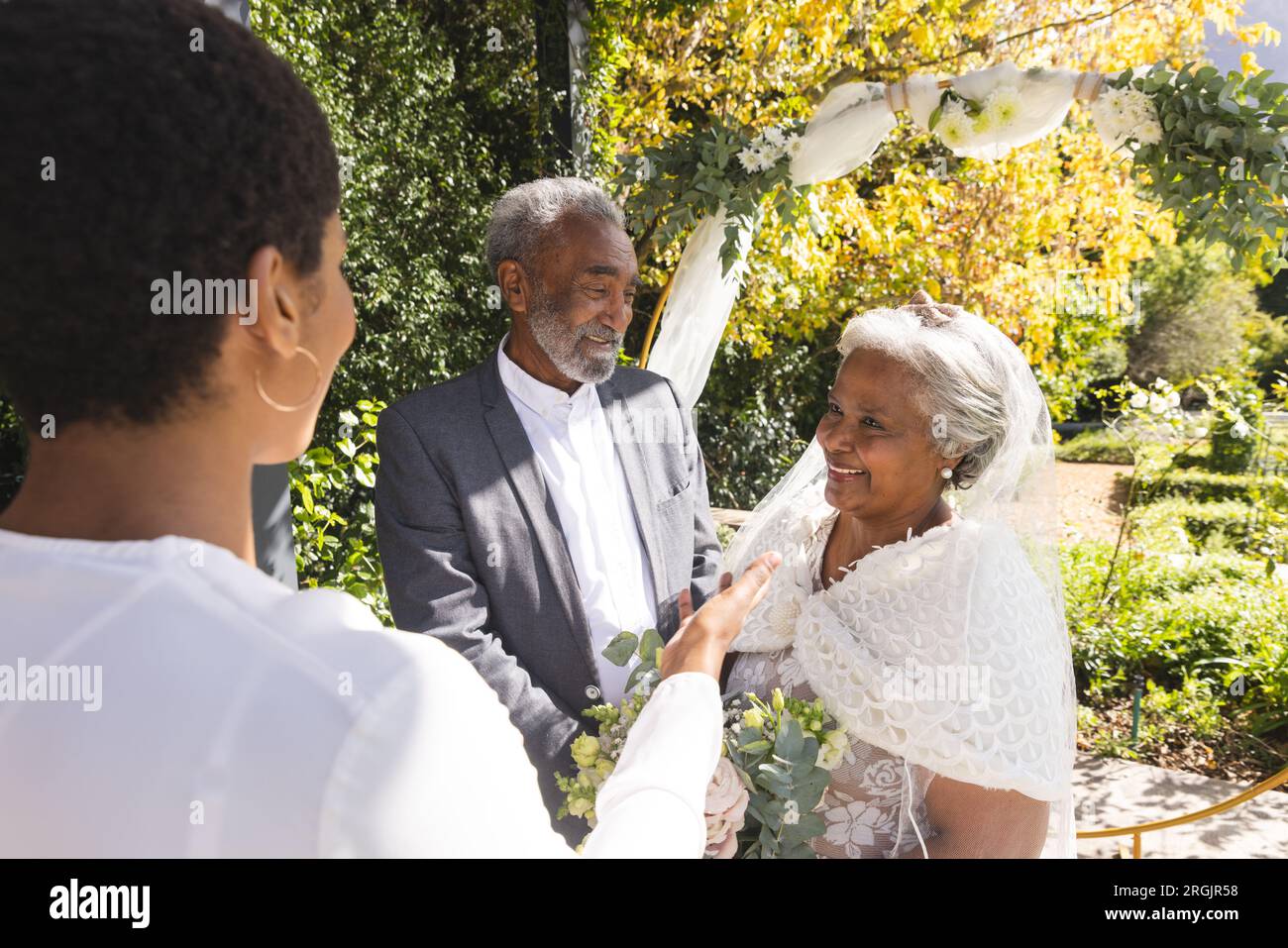Birassische weibliche Ehestandsleiterin und glückliches Seniorenpaar während der Hochzeit im sonnigen Garten Stockfoto