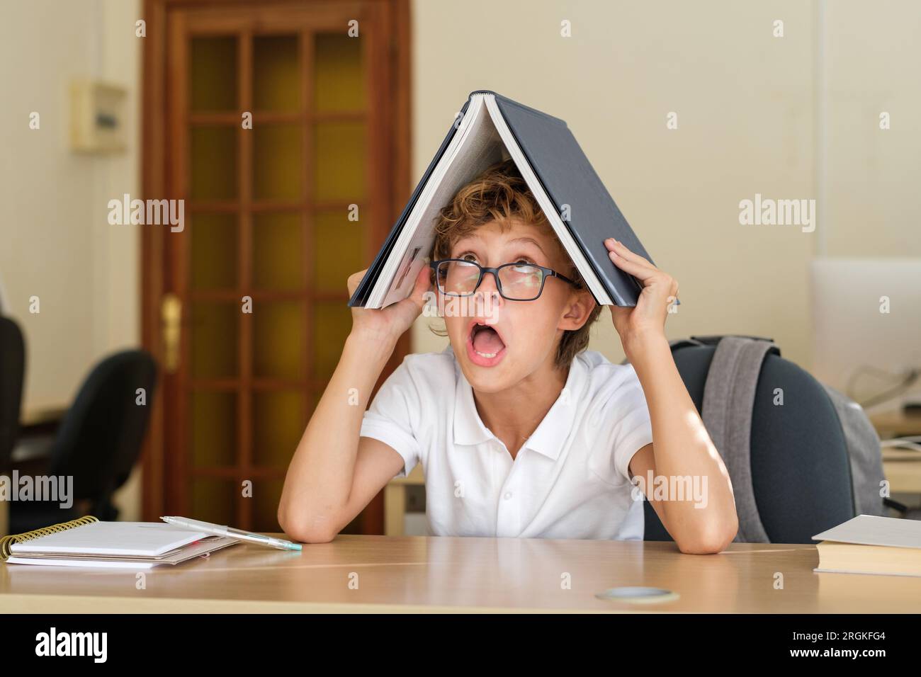 Lustiges Schulkind in einer Brille, das Gesicht macht, während es mit einem Buch auf dem Kopf am Tisch im Klassenzimmer sitzt Stockfoto