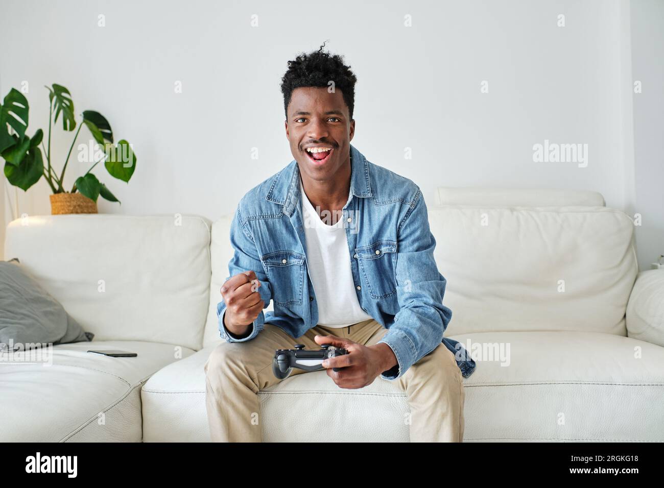 Fröhlicher afroamerikanischer Gamer in lässiger Kleidung, der Videospiel spielt und vor Aufregung die Faust hebt, während er auf einem bequemen Sofa sitzt Stockfoto