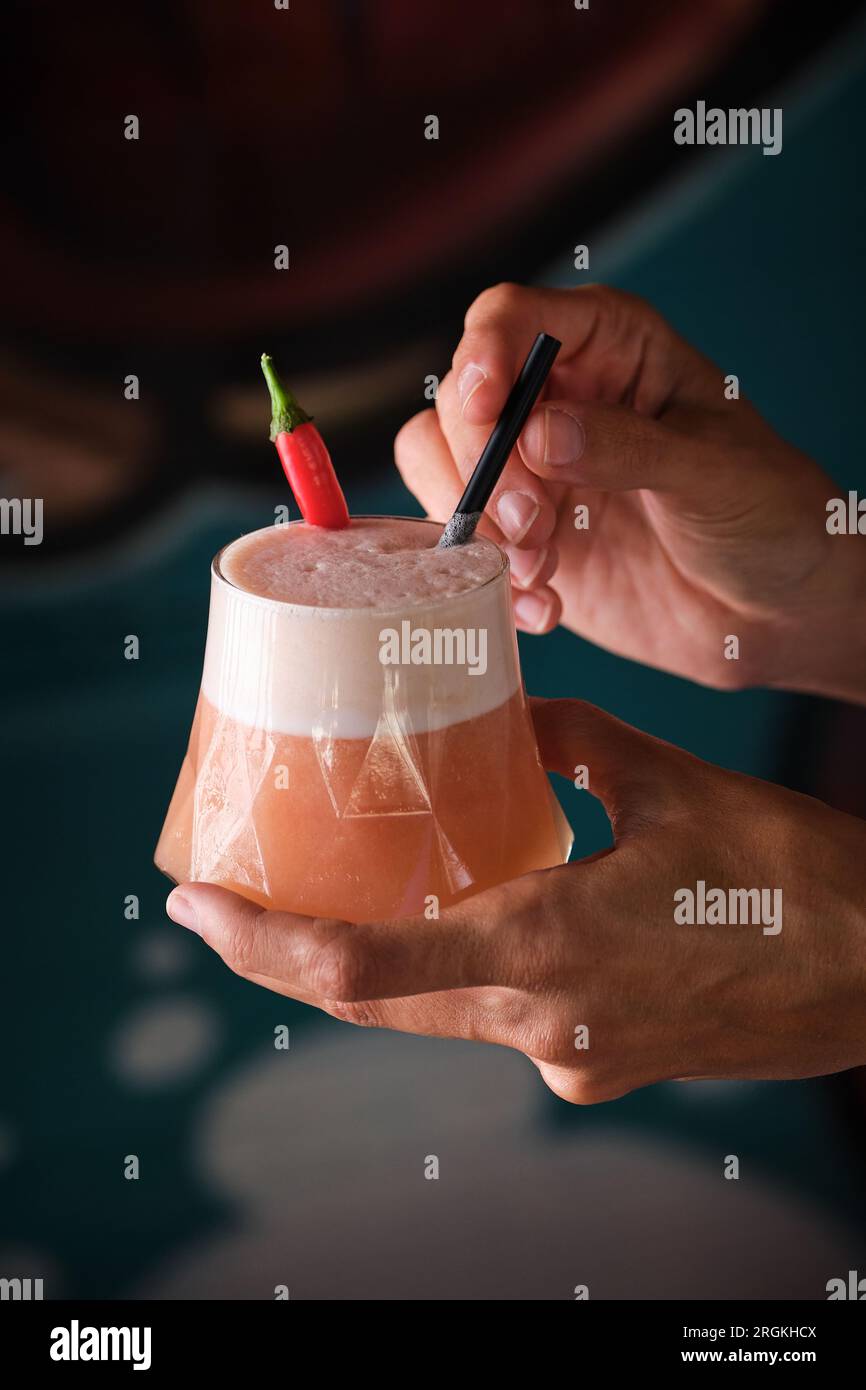 Eine Person, die köstliche Cocktails mit Chili-Pfeffer vorführt und in Glas mit Stroh auf verschwommenem Hintergrund serviert wird Stockfoto