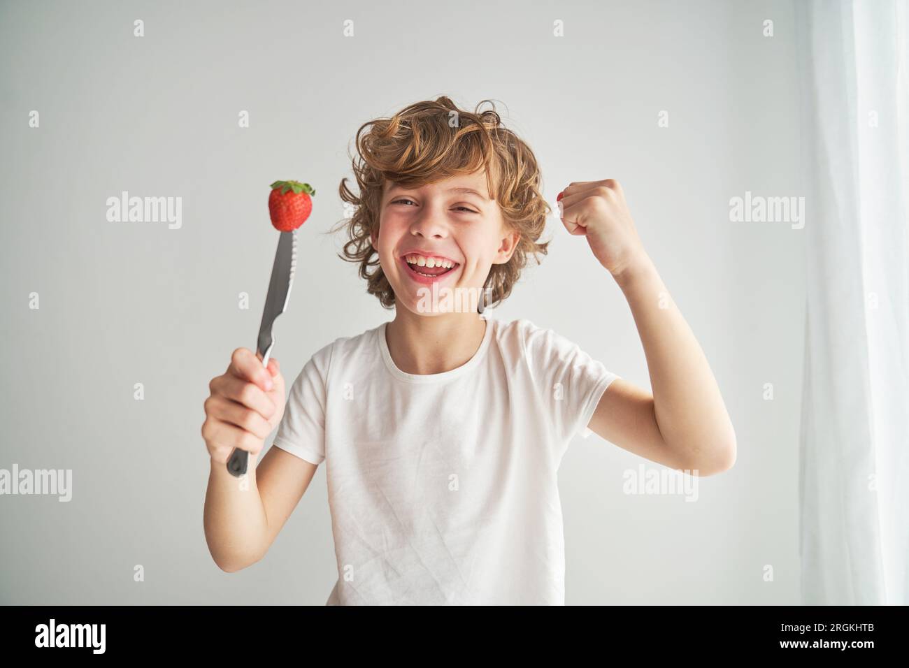 Lächelndes Kind mit frischer Beere, durchbohrt mit Messer, schaut in die Kamera und feiert den Erfolg mit geballter Faust auf weißem Hintergrund Stockfoto