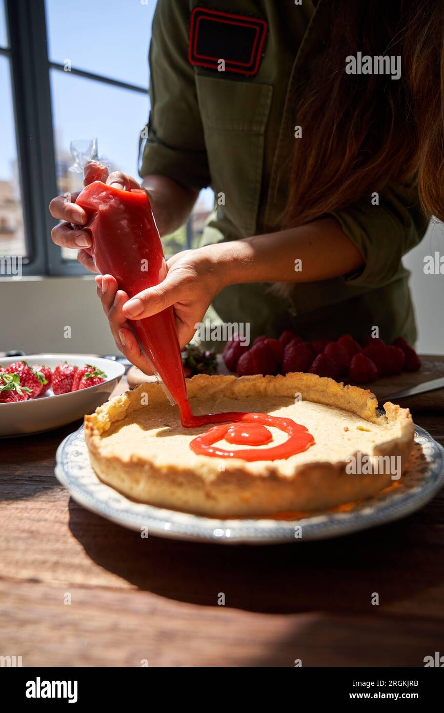 Schneide die weibliche Köchin in lässiger Kleidung, die am Tisch steht, und lege Beerensauce auf die Kuchenkruste, während du leckeren Kuchen zubereitest Stockfoto