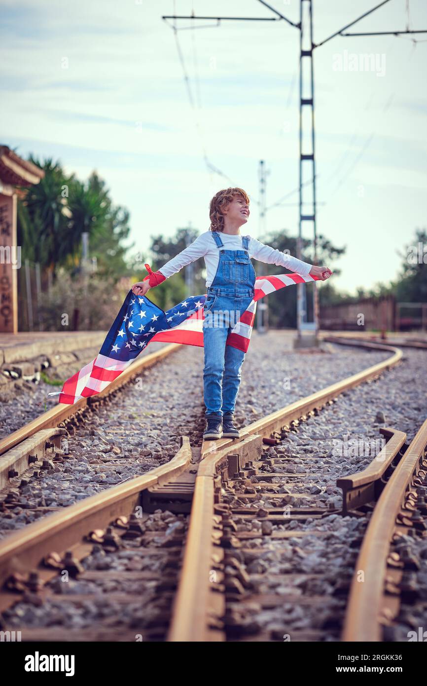 Blondes Kind in Jeans, das auf dem Bahnschalter steht und glücklich wegschaut, während es die flatternde amerikanische Flagge in den Händen hält Stockfoto