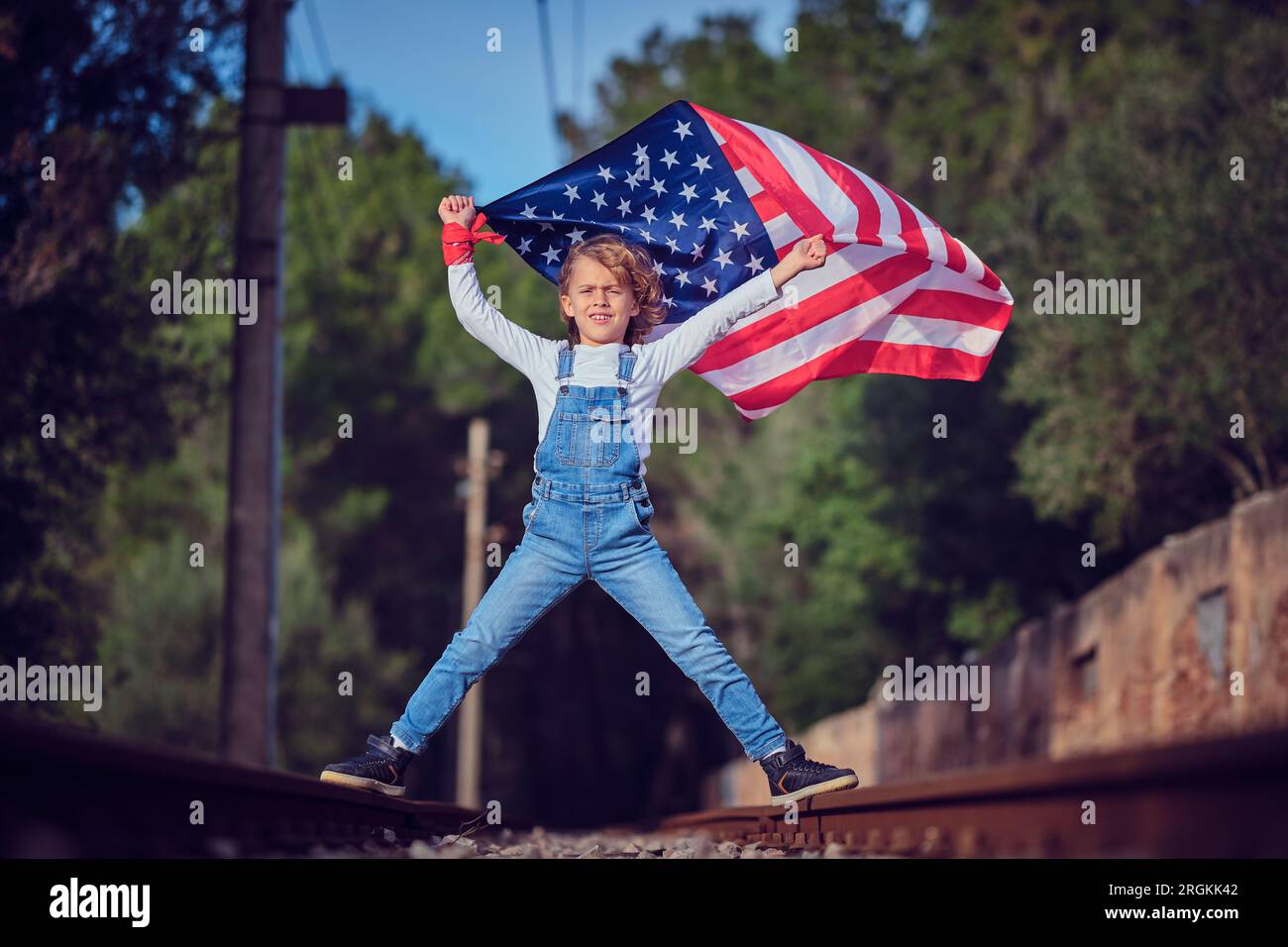 Der ganze Körper eines blonden Jungen, der die flatternde amerikanische Flagge in den Händen hockt und stolz auf die Kamera schaut, während er auf der Eisenbahn steht Stockfoto