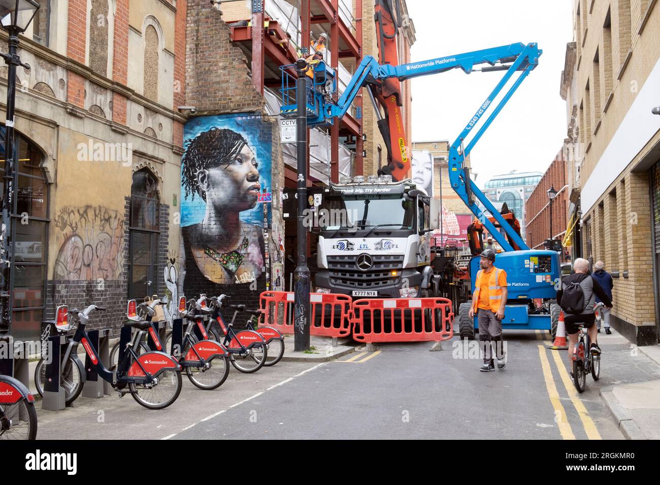 Straße gesperrt durch Bauwagen mit Kran und Cherrypicker Fahrzeuge schwarz Frauengemälde Spitalfields East London E1 UK England KATHY DEWITT Stockfoto