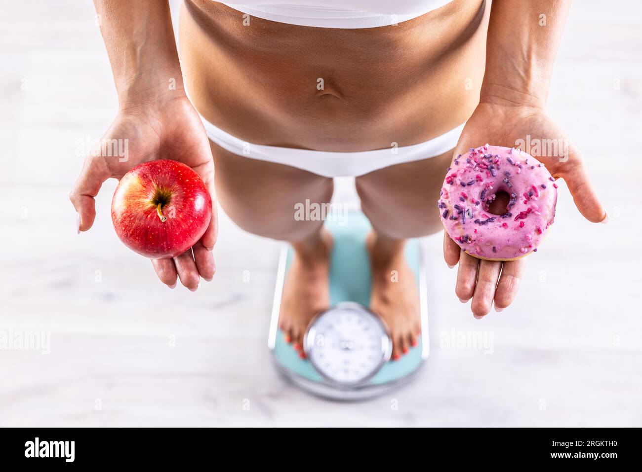 Sportliche Frau mit einem perfekten Körper, der Gewicht auf der Waage misst und einen Donut und einen roten Apfel hält.gesundes oder ungesundes Lifestyle Konzept. Stockfoto