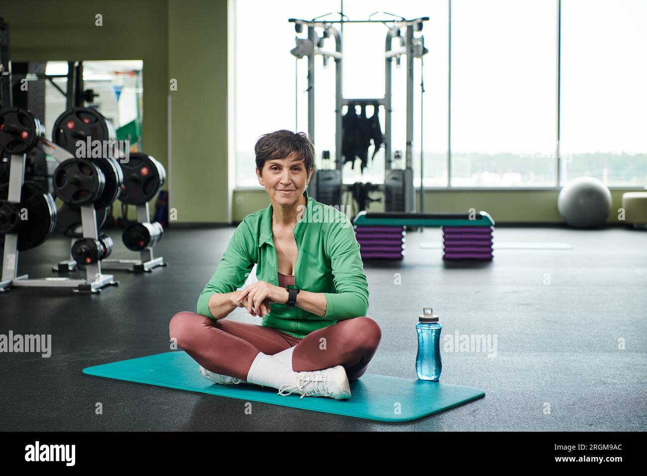 Fröhliche ältere Frau, die mit gekreuzten Beinen auf der Fitnessmatte sitzt, Sportflasche mit Wasser, aktiv Stockfoto
