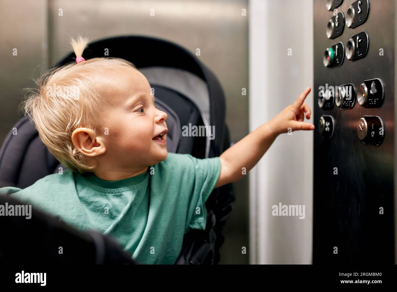 Ein kleines süßes Baby, das in einem Kinderwagen sitzt, drückt den Knopf am Boden im Fahrstuhl. Eine kleine Blondine in einer Kutsche fährt mit ihm den Aufzug hoch oder runter Stockfoto