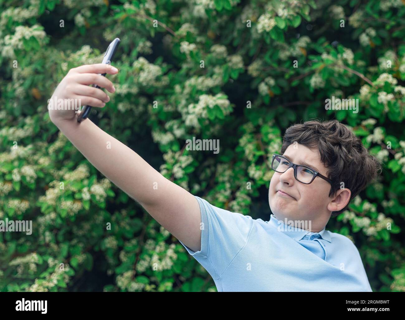 Porträt eines 11-jährigen Jungen in Brille, der Selfie macht, einen Baum oder Busch im Hintergrund blüht Stockfoto