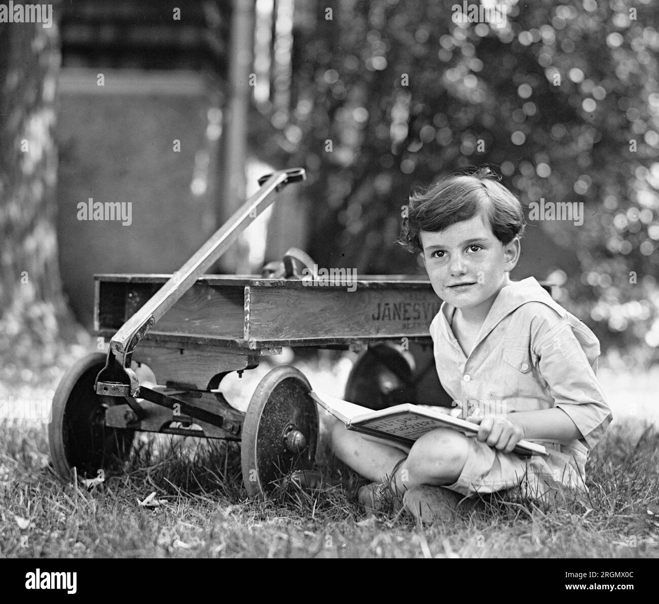 Ein Junge, der in einem Hof neben einem Wagen sitzt und ein Buch liest. 1923 Stockfoto