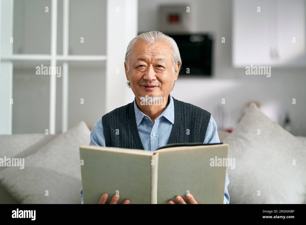 Ein glücklicher alter asiatischer Mann, der zu Hause auf der Couch sitzt und ein Buch liest, in die Kamera schaut und lächelt Stockfoto
