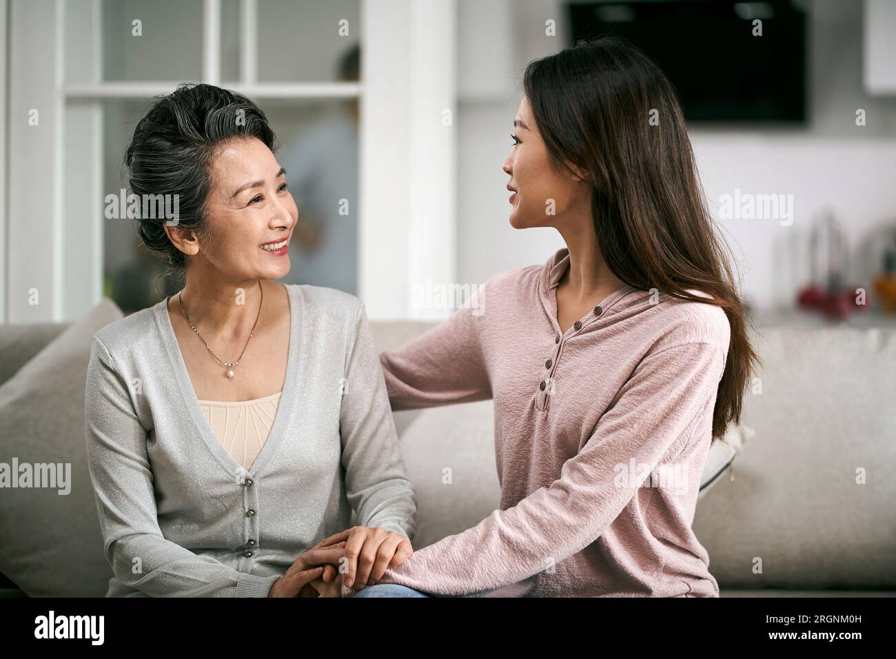 asiatische Erwachsene Tochter, die zu Hause auf der Couch sitzt und sich mit der älteren Mutter unterhält, glücklich und lächelnd Stockfoto