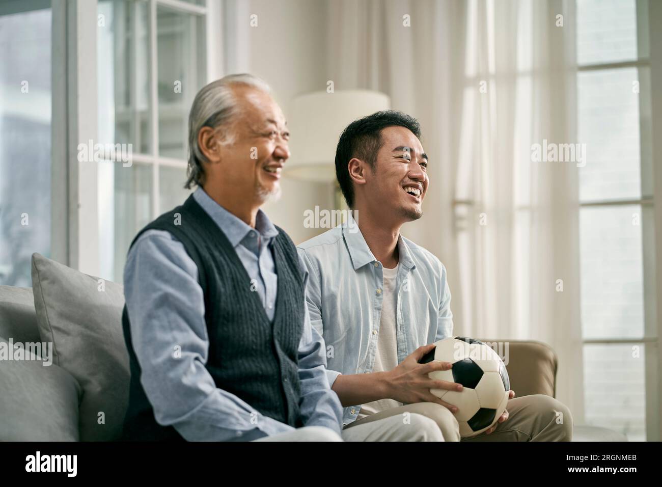 Älterer asiatischer Vater und erwachsener Sohn sitzen zu Hause auf der Couch und sehen sich zusammen Fußballspiele im fernsehen an Stockfoto