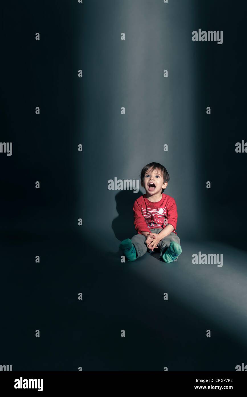 Ein Kleinkind sitzt in einem Lichtstreifen, der durch eine Tür kommt Stockfoto