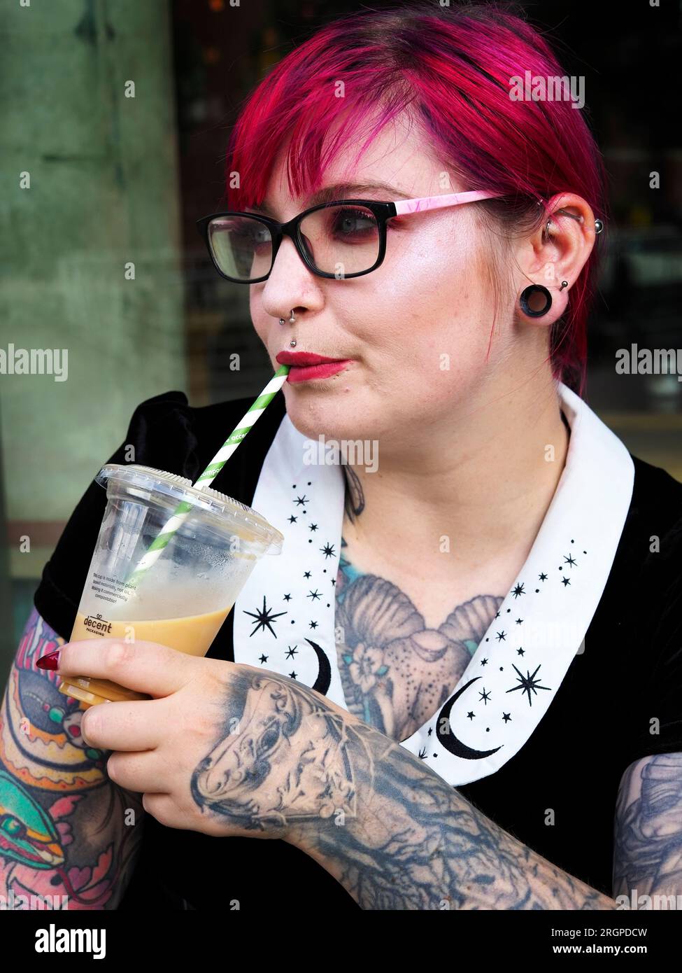 Eine Frau Mitte zwanzig mit gefärbtem rotem Haar und Tätowierungen, die Gläser trägt und vor einem Café in Leeds West Yorkshire, England, Eiskaffee trinkt Stockfoto