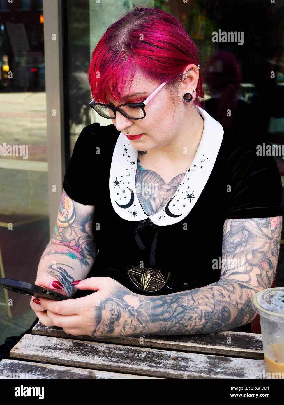 Eine Frau Mitte zwanzig mit gefärbtem rotem Haar und Tätowierungen, die eine Brille mit einem Smartphone trägt, in Leeds West Yorkshire, England Stockfoto