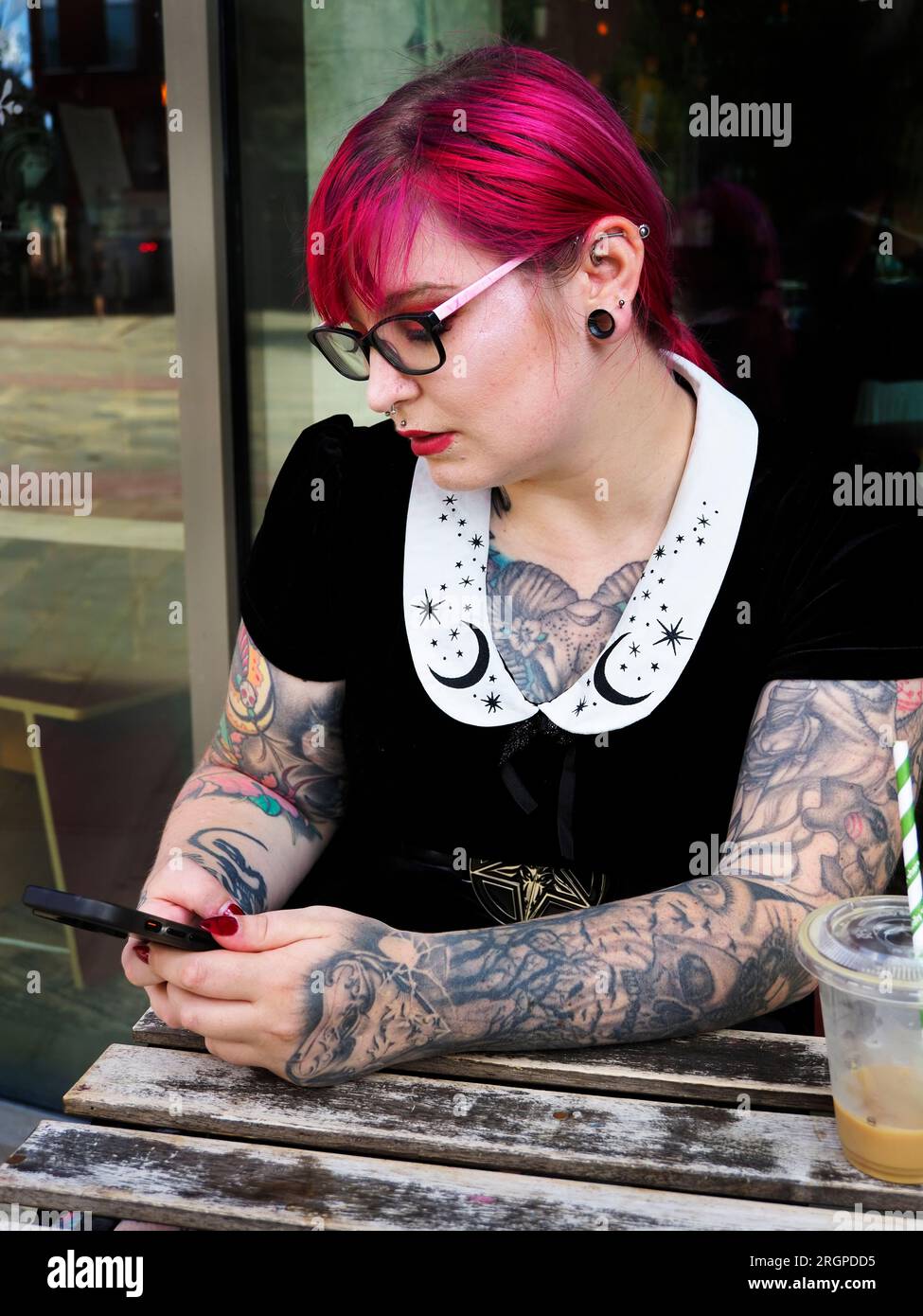 Eine Frau Mitte zwanzig mit gefärbtem rotem Haar und Tätowierungen, die eine Brille mit einem Smartphone trägt, in Leeds West Yorkshire, England Stockfoto