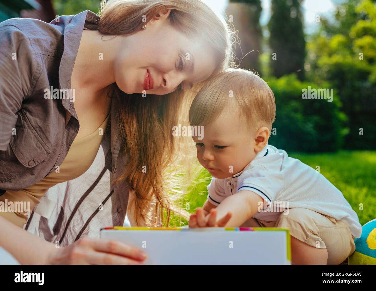 Junge Mutter mit einjährigem Kind, die im Park sitzt und ein Buch liest Stockfoto