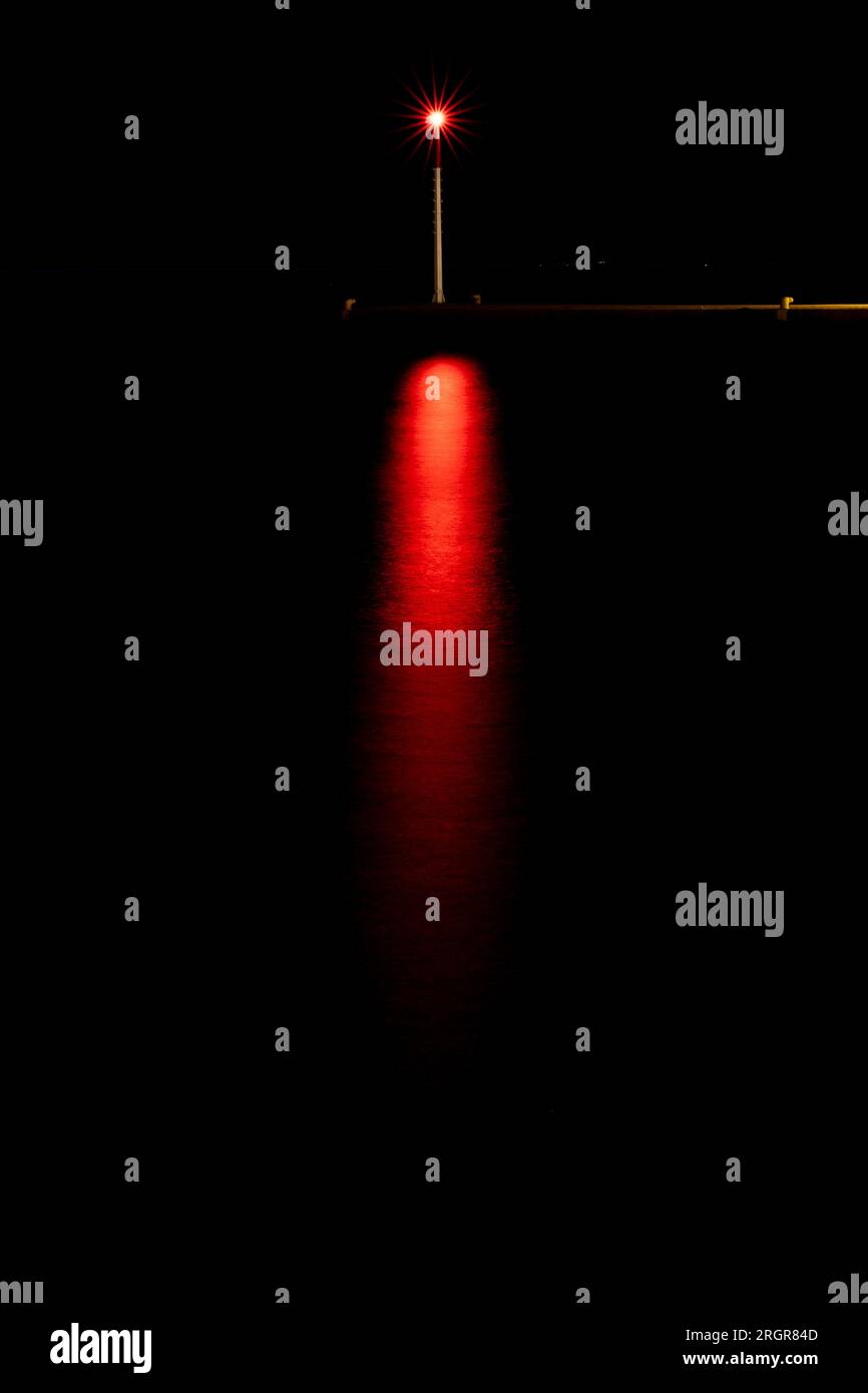 Langzeitbelichtungs-Foto einer Straßenlampe am Dock der Bucht von Spetses mit rotem Licht bei Nacht vor schwarzem Himmel mit Reflexion auf Meerwasser Stockfoto