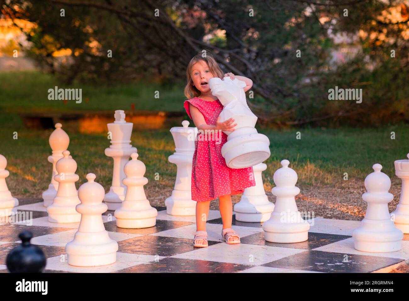 Kleines Weißes Mädchen in einem rosa Kleid, das Straßenschach spielt. An einem Sommerabend spielt ein Kind im Park mit großen Schachfiguren. Stockfoto