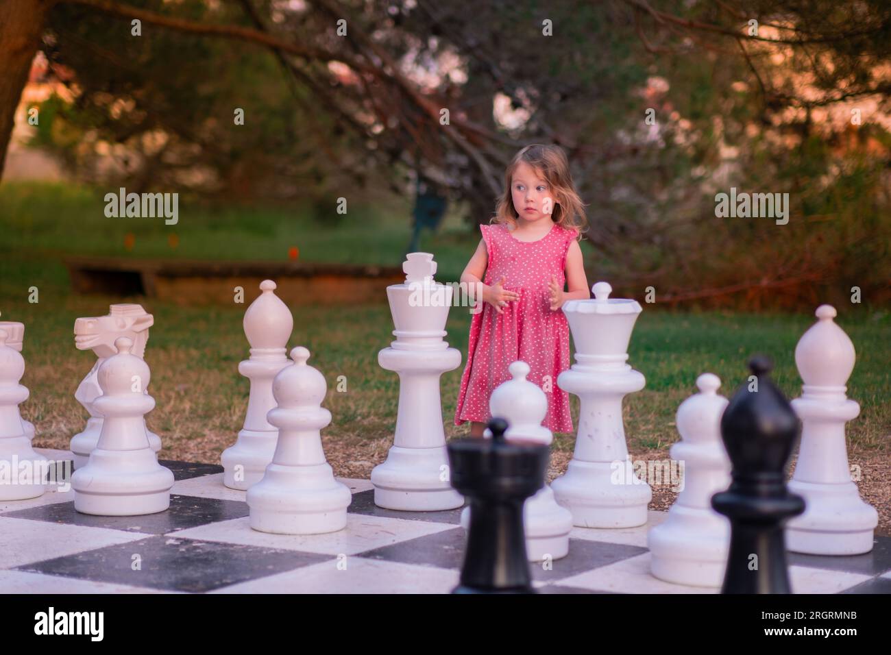 Kleines Weißes Mädchen in einem rosa Kleid, das Straßenschach spielt. An einem Sommerabend spielt ein Kind im Park mit großen Schachfiguren. Stockfoto