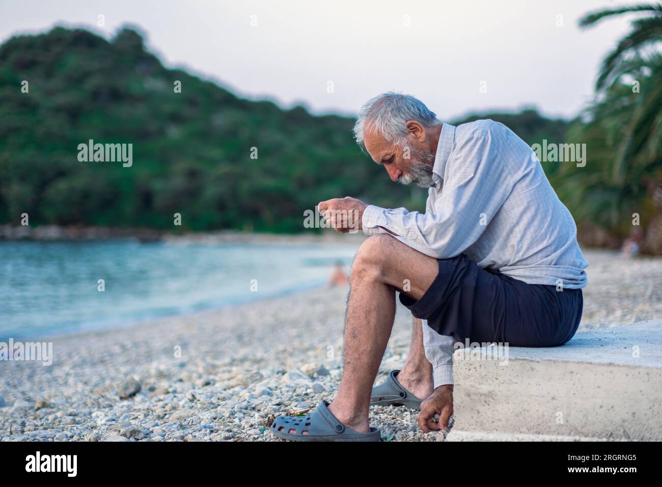 Ein alter Mann am Strand in einem weißen Hemd und dunklen Hosen. Ein alter Mann mit Bart geht bei Sonnenuntergang allein am Strand spazieren. Porträt eines einsamen Grauhaars Stockfoto