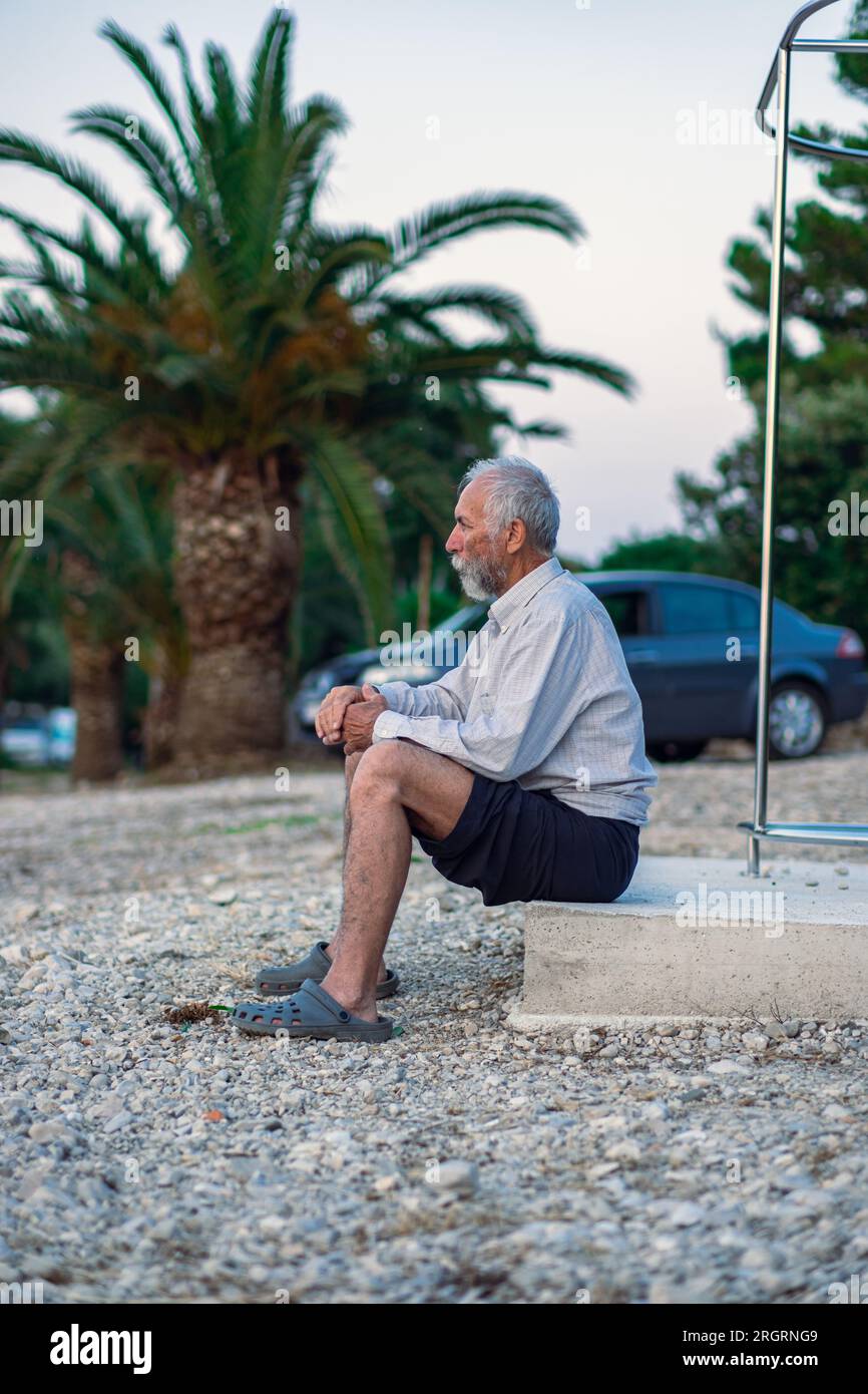 Ein alter Mann am Strand in einem weißen Hemd und dunklen Hosen. Ein alter Mann mit Bart geht bei Sonnenuntergang allein am Strand spazieren. Porträt eines einsamen Grauhaars Stockfoto