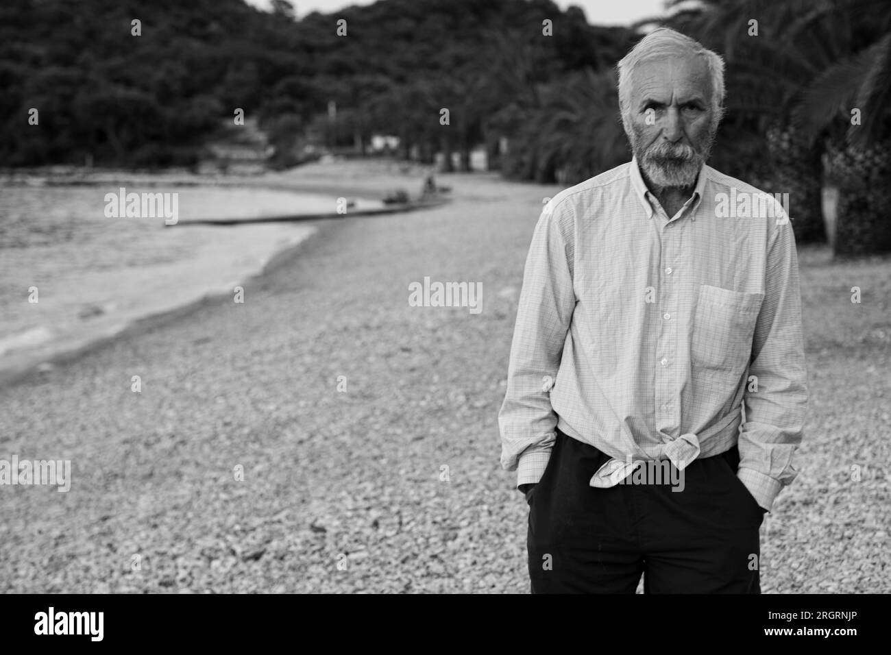 Ein alter Mann am Strand in einem weißen Hemd und dunklen Hosen. Ein alter Mann mit Bart geht