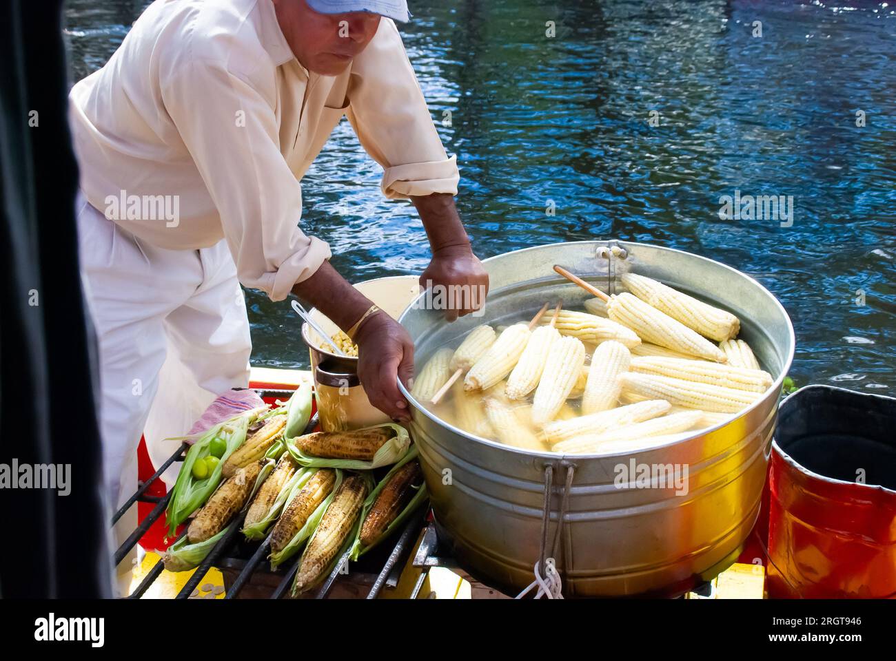 Ein Verkäufer bereitet gekochten und gerösteten Mais geschickt auf einem Boot in Xochimilco, Mexiko, zu und verkauft ihn. Die große Wanne mit dampfenden Ähren aus Mais fügt ein hinzu Stockfoto