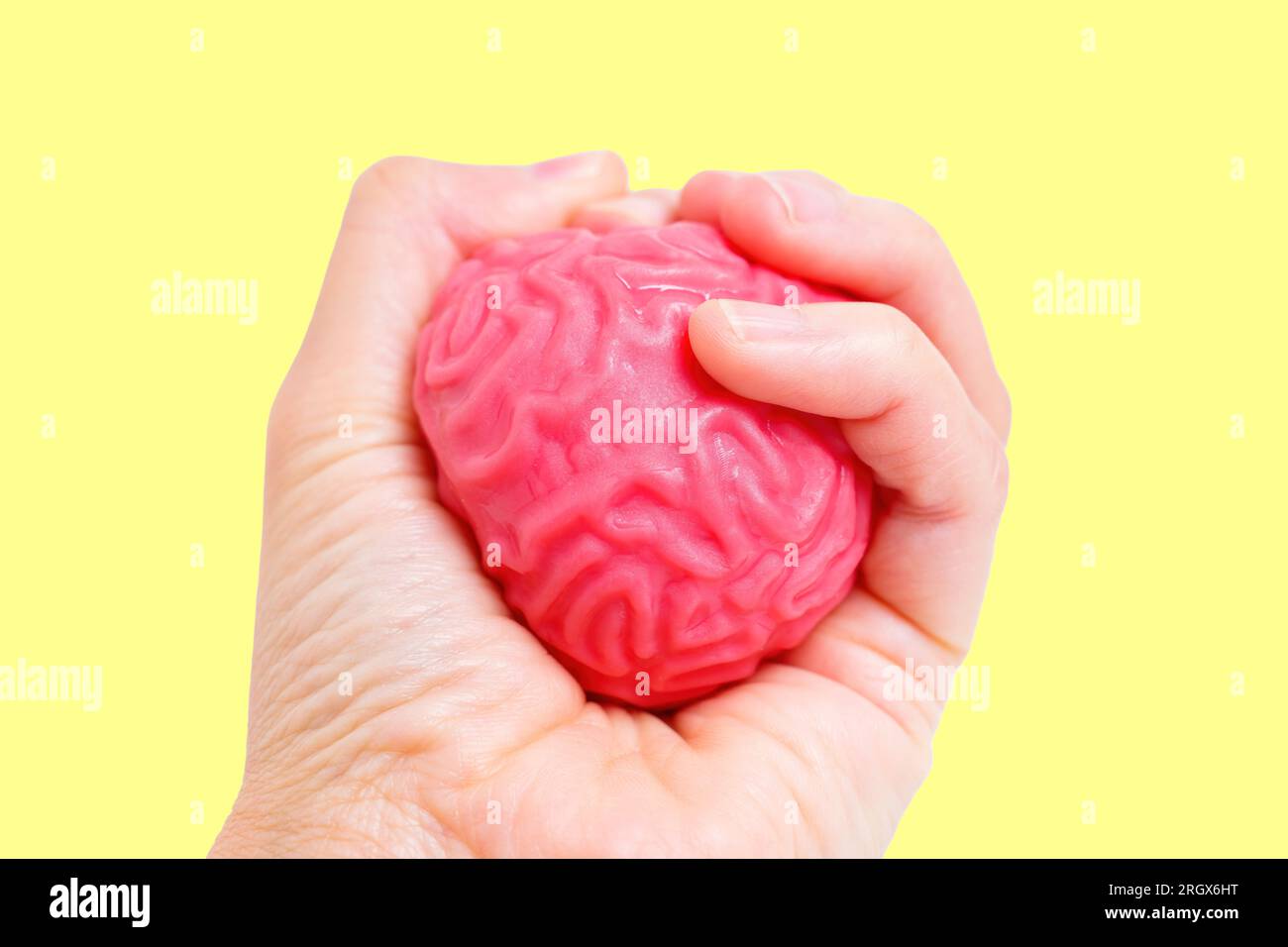 Hand demonstriert Kontrolle und Einfluss, indem sie ein geschmeidiges, gelbähnliches menschliches Hirnmodell fest zusammendrückt, isoliert auf gelbem Hintergrund. Kognitives ab Formen Stockfoto
