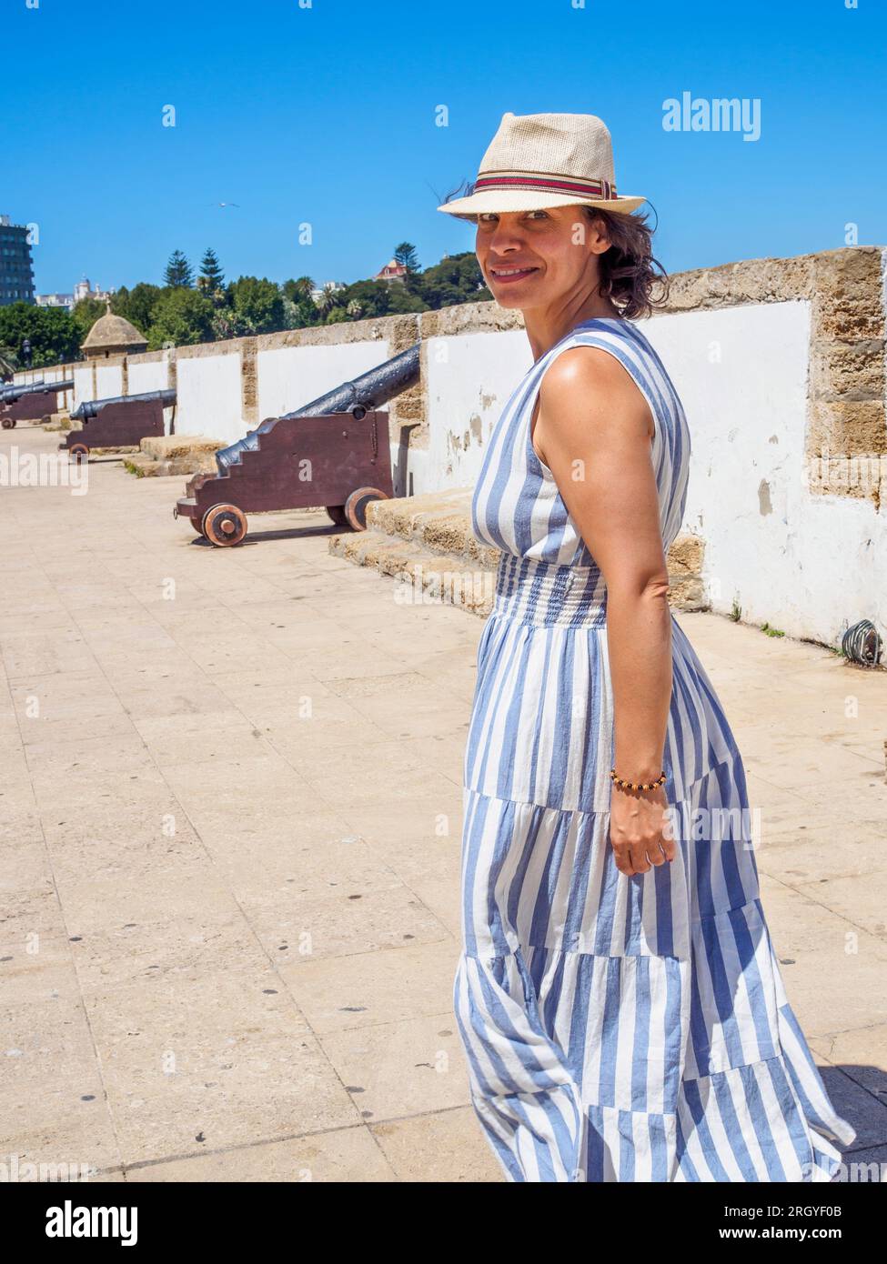 Lächelnder kaukasischer Tourist, der ein Sommerkleid und einen alten Strohhut trägt und auf einer antiken Latinamerikanischen Yachthafenpromenade spaziert. Stockfoto
