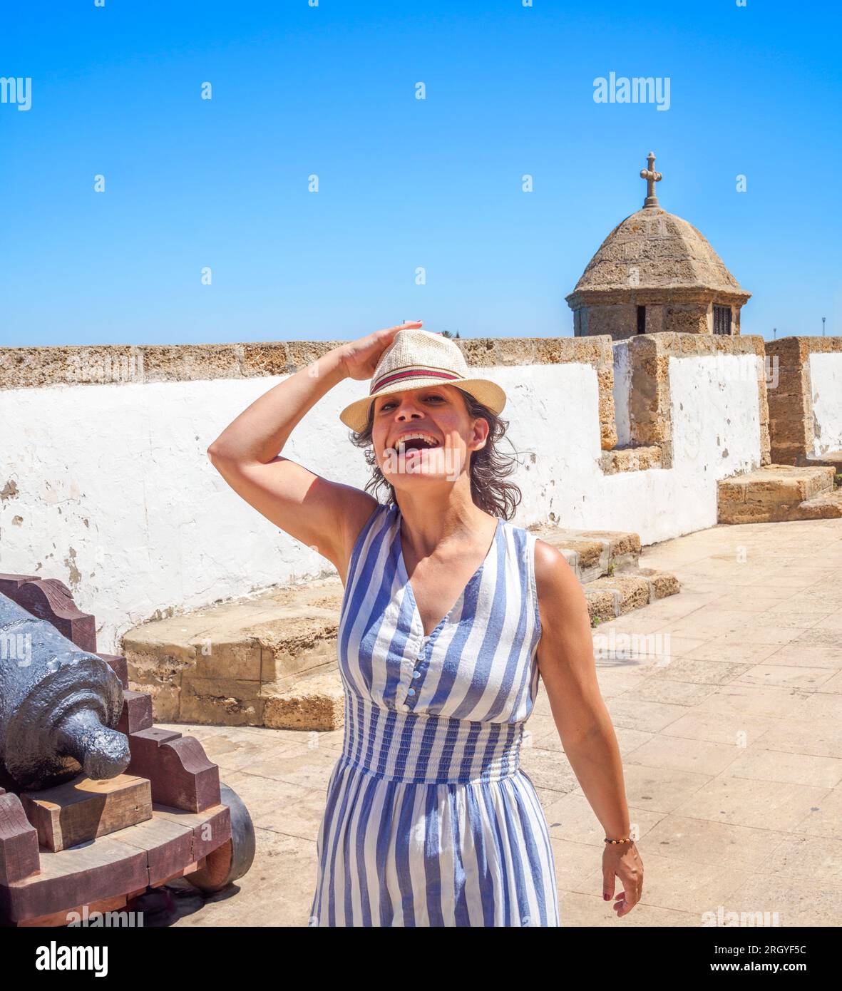 Lächelnder kaukasischer Tourist, der ein Sommerkleid und einen alten Strohhut trägt und auf einer antiken Latinamerikanischen Yachthafenpromenade spaziert. Stockfoto