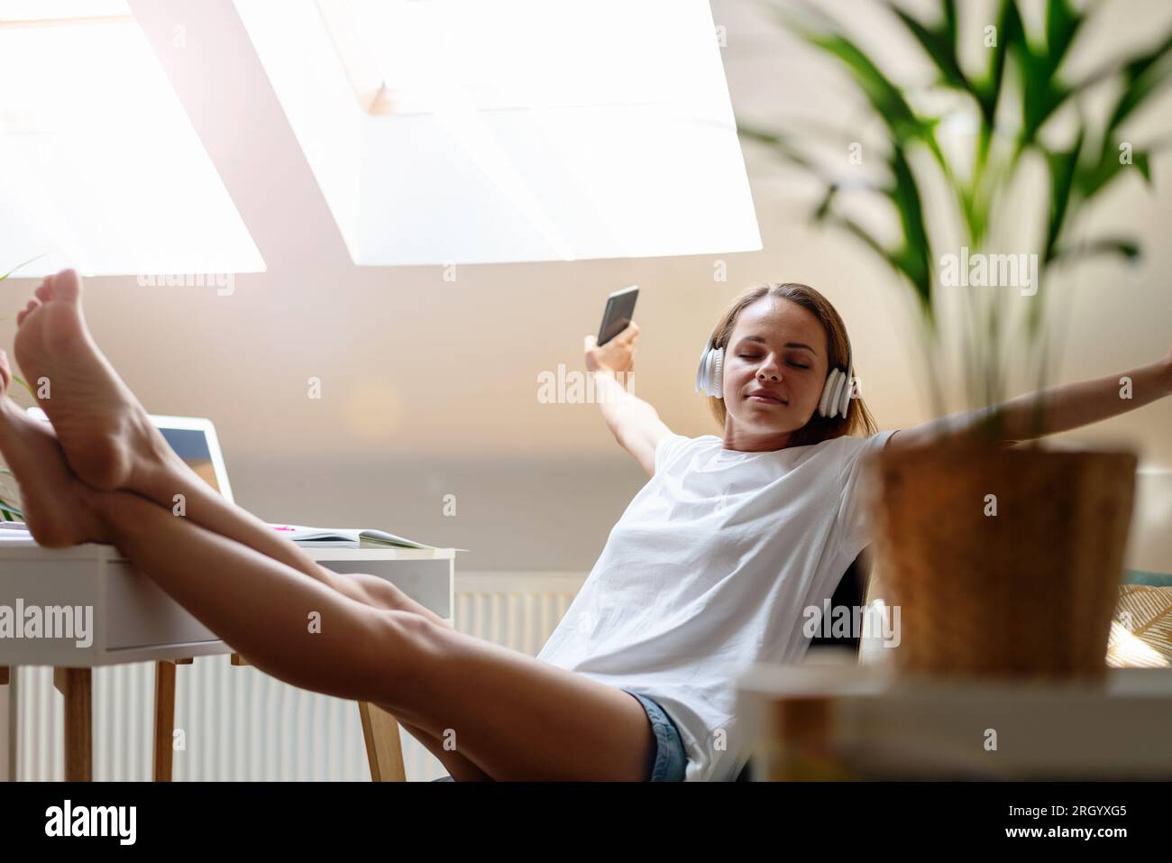 Die junge Frau sitzt mit den Füßen oben auf dem Tisch und hört Musik mit ihren kabellosen Kopfhörern und ihrem Smartphone, genießt Musik, streckt ihre Arme und bo Stockfoto