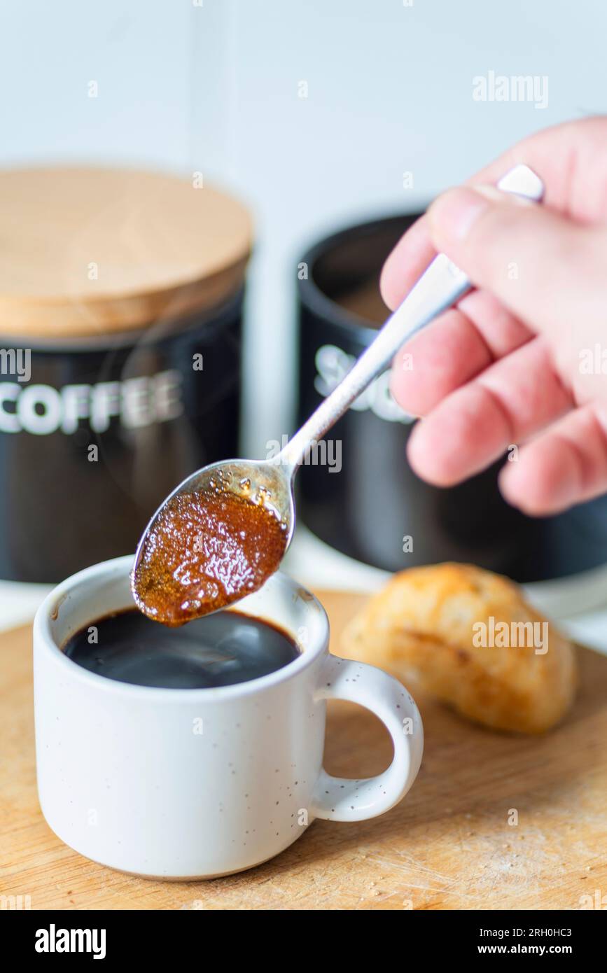 Braune Zuckerkörner ziehen den heißen Kaffee zu einer sirupen Konsistenz auf, bevor sie in einen frisch gebrühten schwarzen Espresso gerührt werden, der trinkfertig und heiß ist Stockfoto