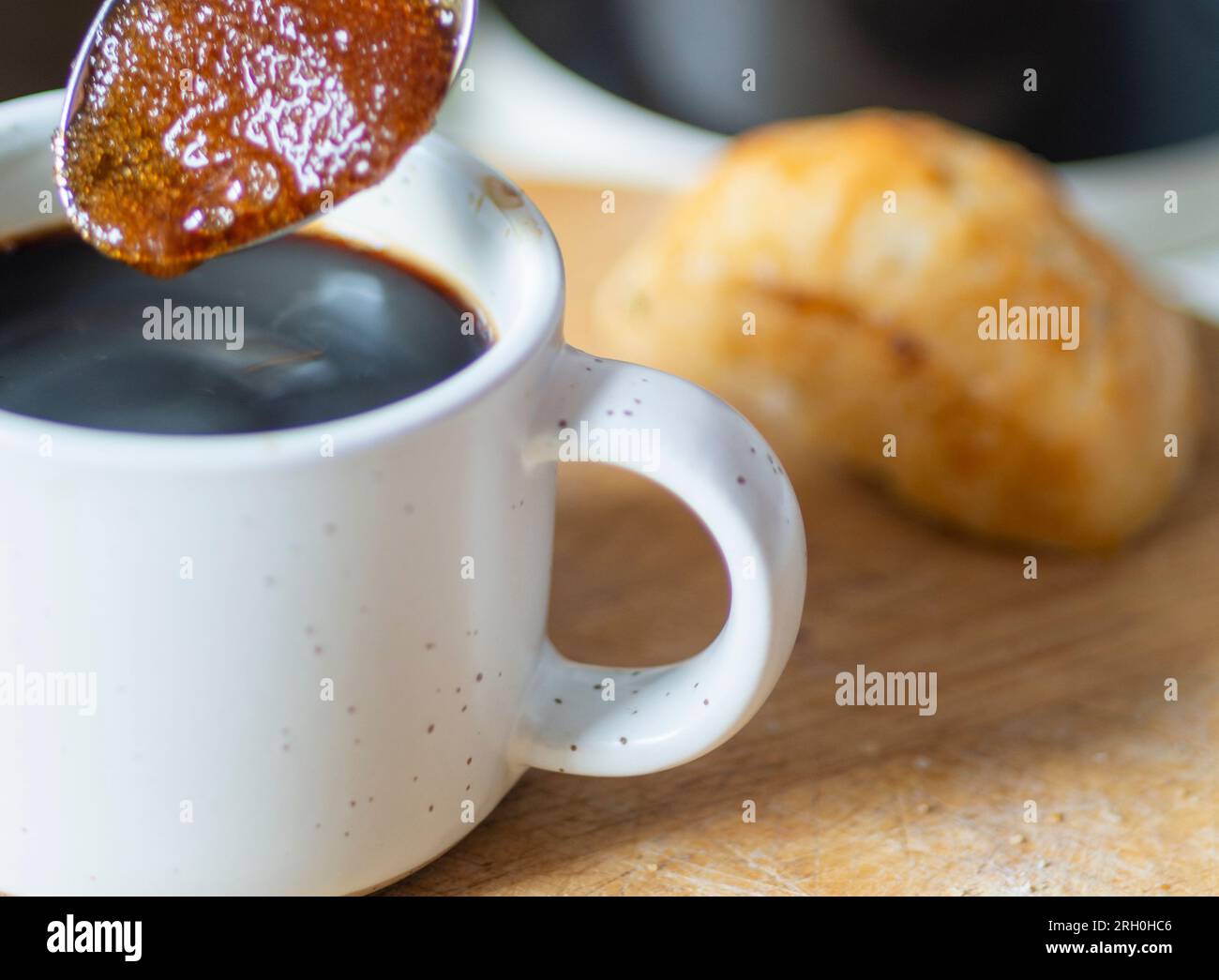 Braune Zuckerkörner ziehen den heißen Kaffee zu einer sirupen Konsistenz auf, bevor sie in einen frisch gebrühten schwarzen Espresso gerührt werden, der trinkfertig und heiß ist Stockfoto