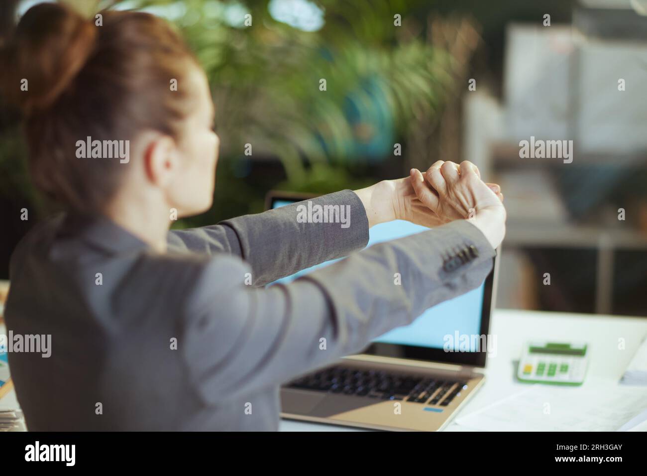 Nachhaltiger Arbeitsplatz. Von hinten gesehen: Moderne Geschäftsfrau in einem grauen Business-Anzug in einem modernen grünen Büro, die sich die Hand streckt. Stockfoto