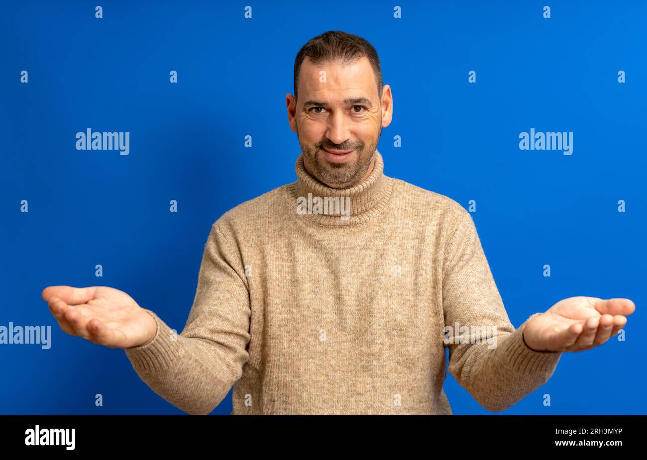 Bärtiger hispanischer Mann in seinen 40s Jahren mit blauem Hintergrund, der in die Kamera schaut und mit offenen Armen lächelt, um sich zu umarmen. Fröhlicher Ausdruck, der Glück umarmt Stockfoto