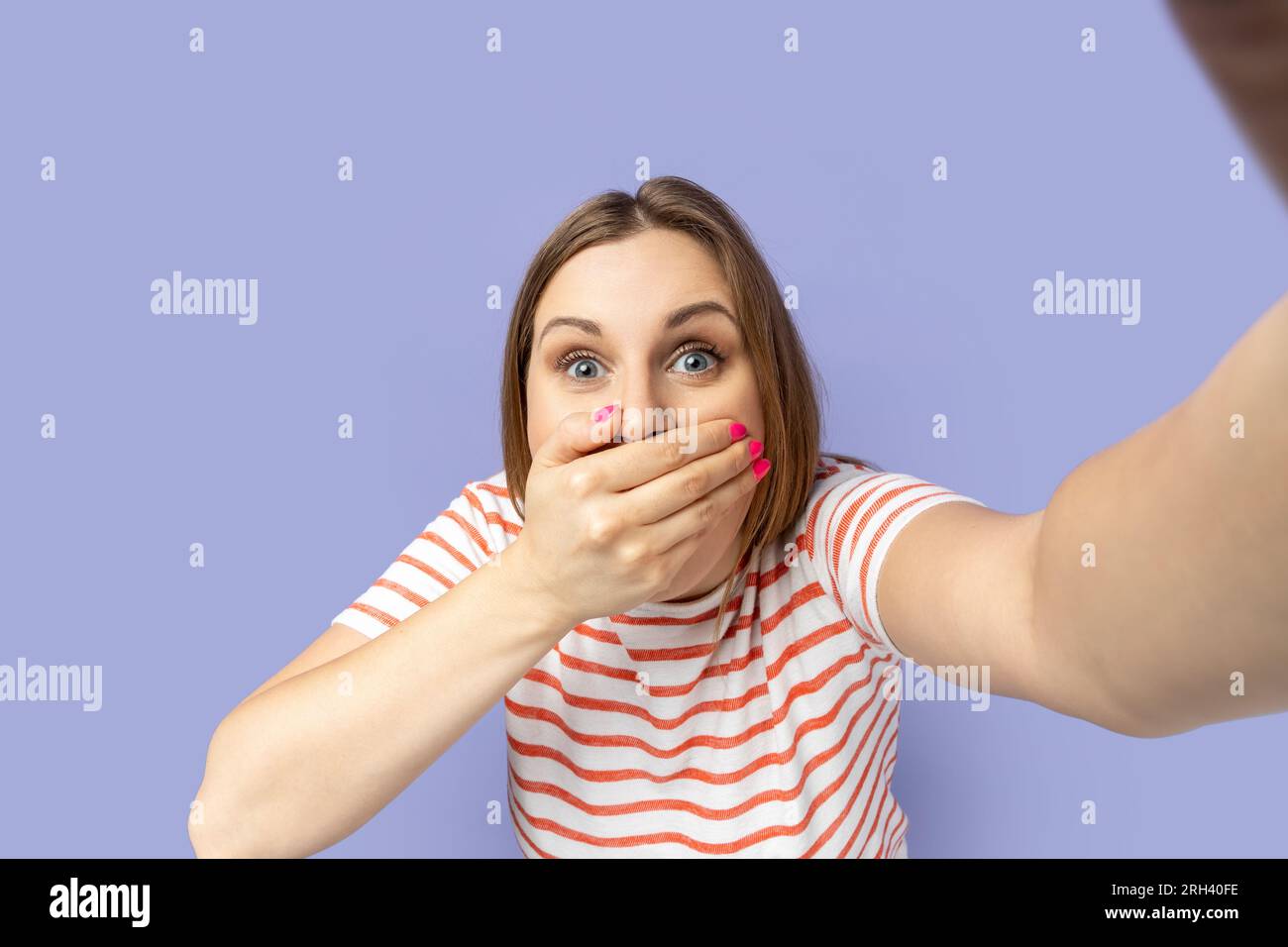 Porträt einer überraschten Frau in gestreiftem T-Shirt, die ein Selfie-Foto macht, ihren Mund mit einer Handfläche bedeckt, Blickwinkel, vorderes Selbstporträt. Studio-Aufnahme im Innenbereich isoliert auf lila Hintergrund. Stockfoto