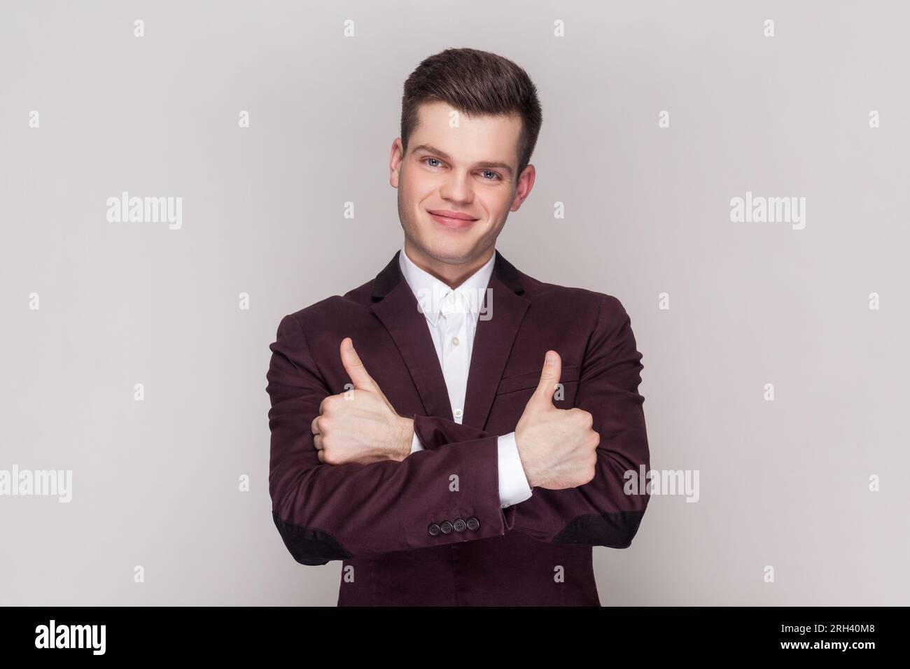 Der Mann macht ein Daumen-hoch-Zeichen, zeigt Unterstützung und Respekt für jemanden, lächelt angenehm vor der Kamera, erreicht das gewünschte Ziel, trägt Anzug und Hemd. Aufnahme im Studio isoliert auf grauem Hintergrund. Stockfoto