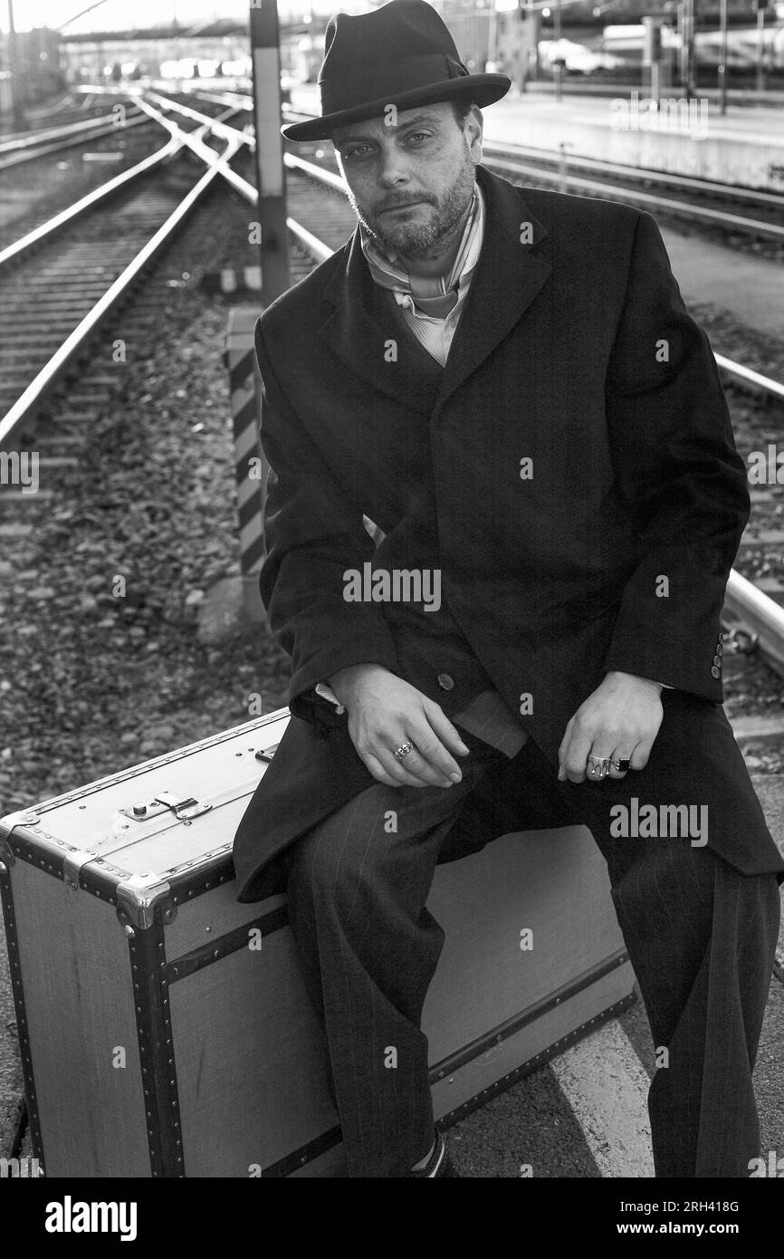 Männlicher Reisender, der auf den Bahnsteig wartet. Mann sitzt am Hauptbahnhof auf Koffern, Zug pünktlich oder verspätet. Stockfoto