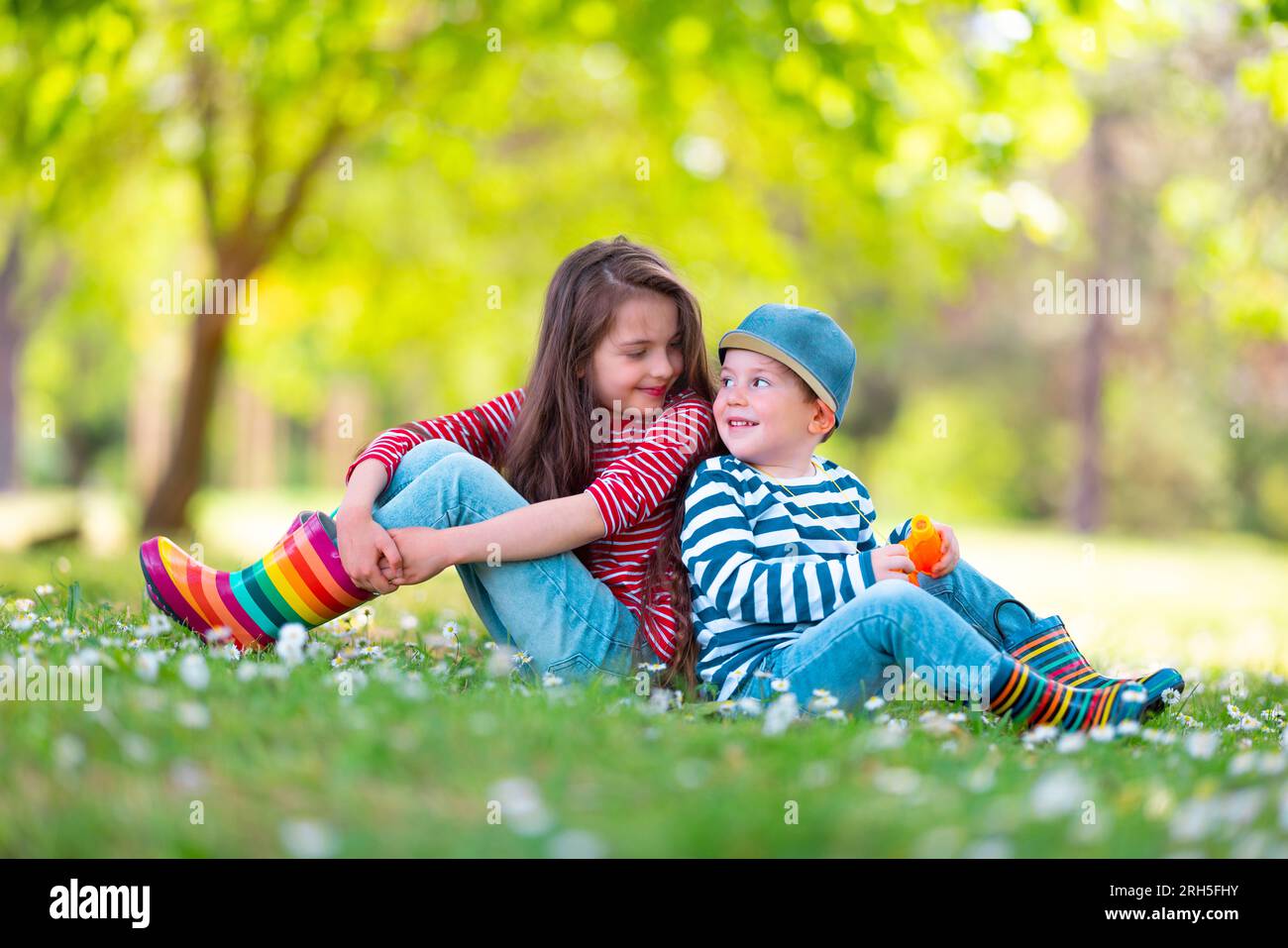 Fröhliche Kinder Junge und Mädchen in Gummistiefeln spielen draußen im grünen Park mit blühenden Blüten Stockfoto