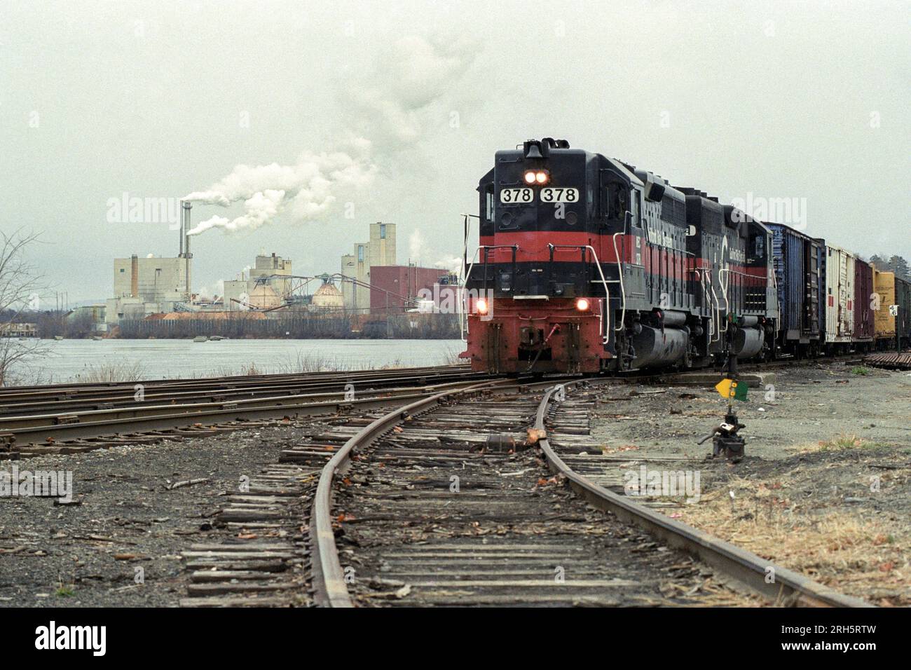 Ein Güterzug fährt an einem bedeckten Tag an einer Papierfabrik vorbei. Stockfoto
