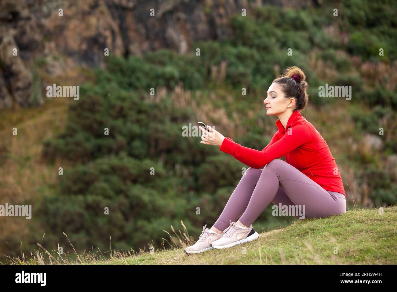 Eine junge Frau, die draußen in Sportbekleidung sitzt und ihr Handy überprüft Stockfoto