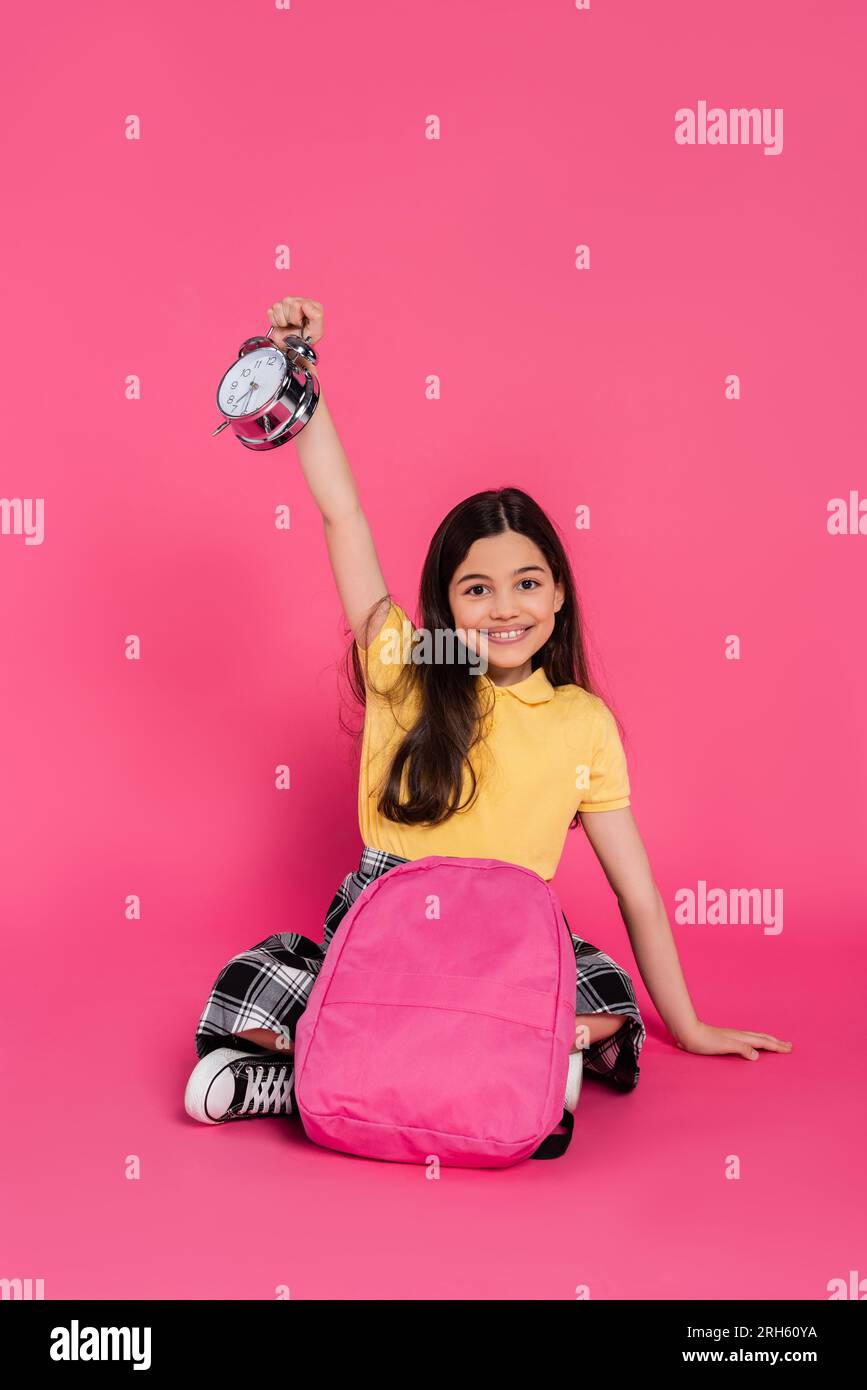 Positives und brünettes Schulmädchen, das mit Rucksack sitzt und den Vintage-Wecker auf Pink hält Stockfoto