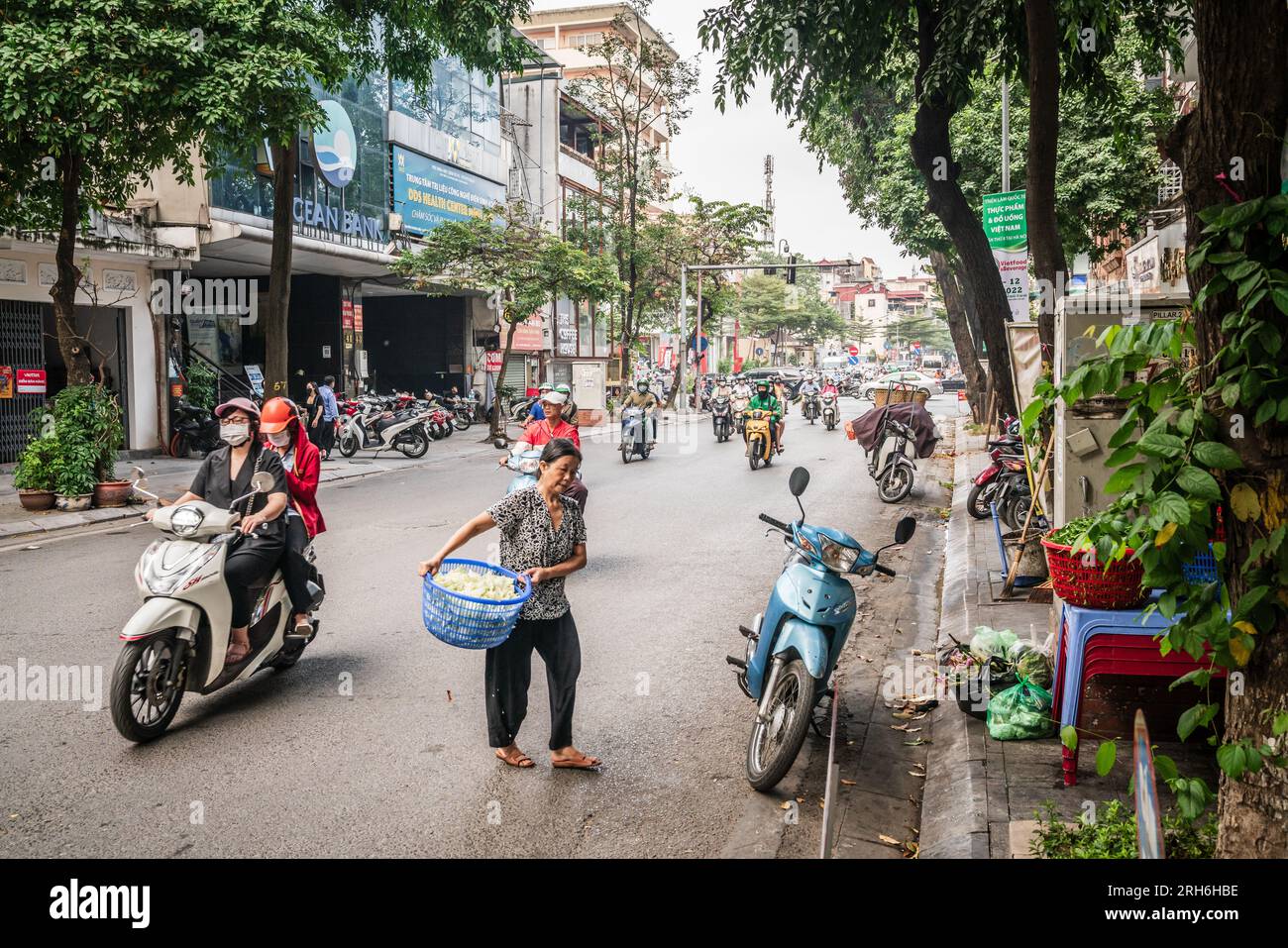 Hanoi, Vietnam, 13. November 2022: Eine ältere Frau mit einem Gemüsekorb navigiert den Rollerverkehr, während sie eine belebte Seitenstraße in Han überquert Stockfoto