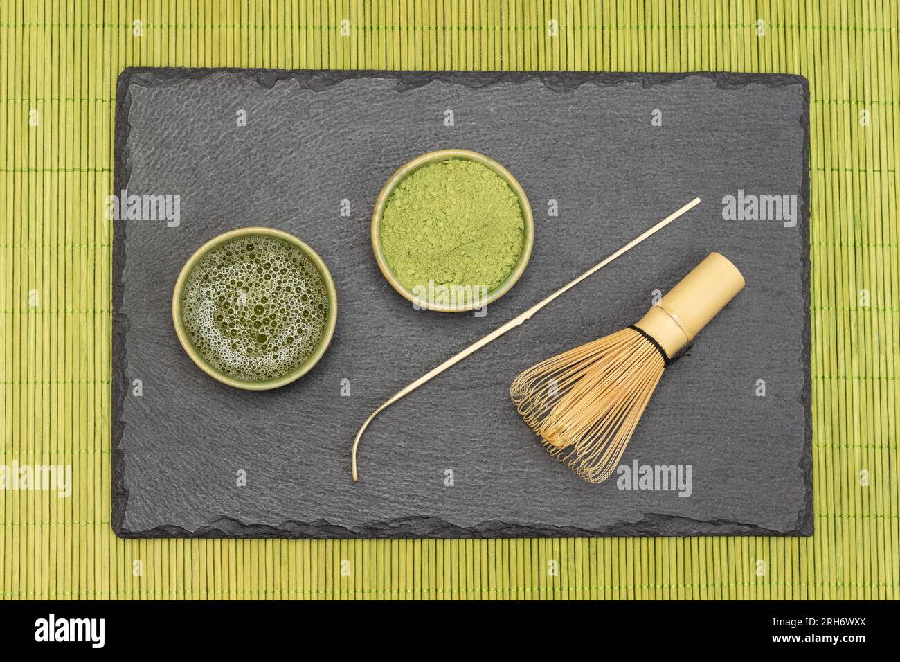 Matcha-Tee und Matcha-Teepulver in Schüssel, Bambusschläger und Teelöffel auf Steinbrett. Grüne Serviette aus Holzstäbchen. Flach verlegt Stockfoto