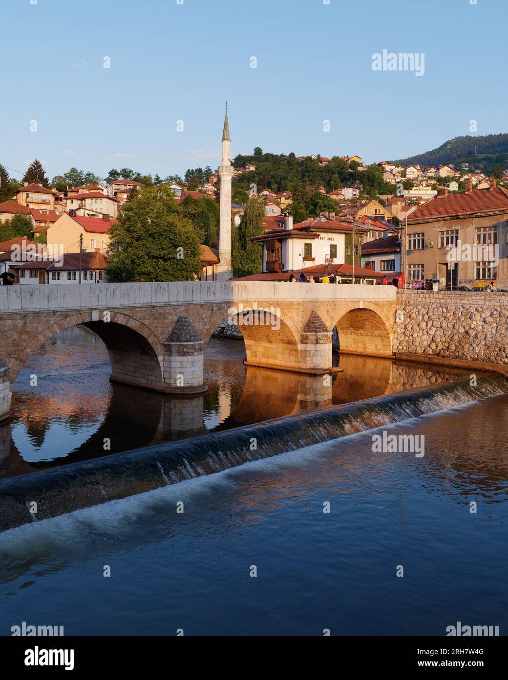 Brücke über den Fluss Miljacka mit Restaurant Inat Kuca und Minarett dahinter in Sarajevo, Bosnien und Herzegowina, 14. August 2023. Stockfoto