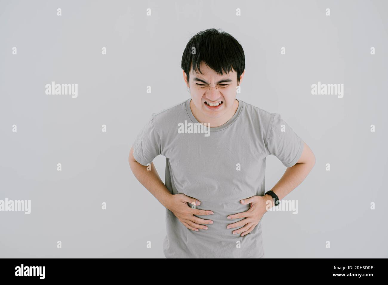 Selektive Fokussierung von einem jungen asiatischen Mann, der ein salbeigrünes lässiges T-Shirt und Smartwatch trug, hatte Bauchschmerzen und hielt ihn mit den Händen an isoliertem weißem Ba Stockfoto